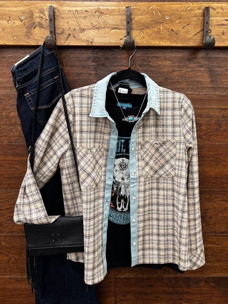 A plaid button-up shirt in beige, brown, and light blue hangs on a black hanger against a rustic wooden wall, styled with dark blue jeans, a black crossbody bag, and a black graphic t-shirt.