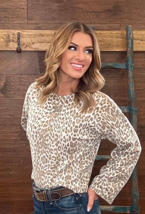 A woman wearing a beige and brown leopard print long-sleeve top with blue jeans, posing in front of a rustic wooden wall with a ladder.