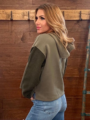 A woman wearing an olive green hooded sweatshirt with contrasting dark green ribbed sleeves, paired with blue jeans, standing in front of a rustic wooden wall.