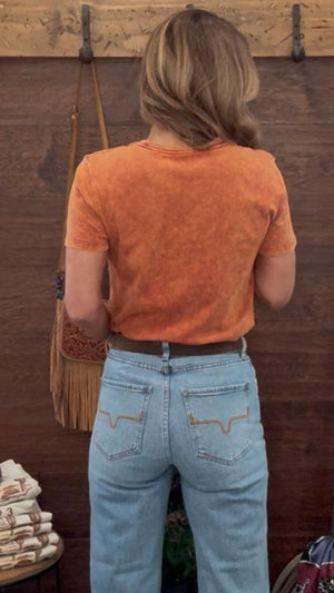 Person wearing an orange shirt and light blue jeans standing in a room with wooden walls.
