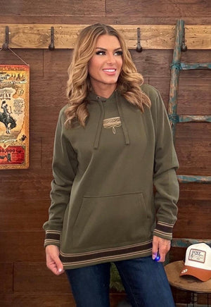 A woman wearing an olive green pullover hoodie with striped cuffs and hem, standing in front of a rustic wooden wall with western decor.