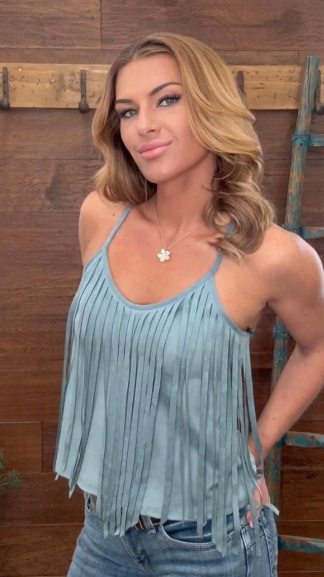 Woman wearing a light blue fringe top and jeans standing in front of a wooden wall with a ladder.