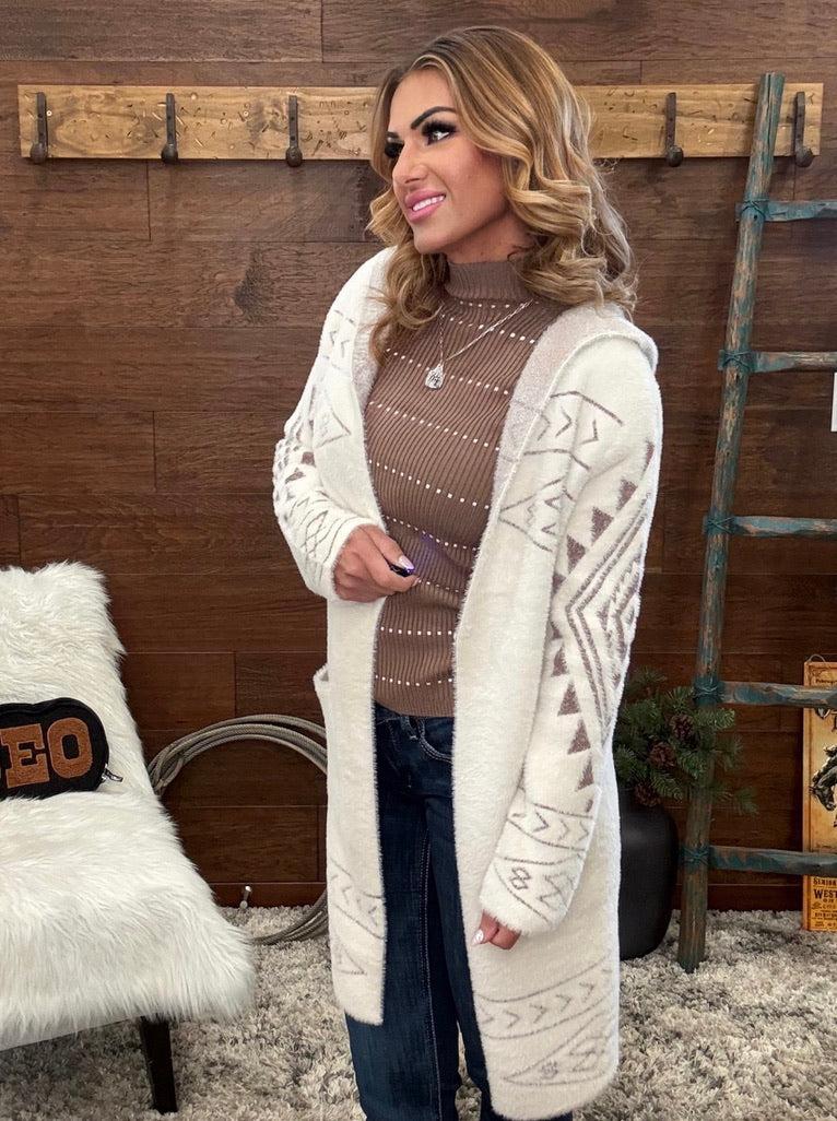 A woman wearing a long cream-colored fuzzy cardigan with brown geometric patterns, styled over a brown knit turtleneck sweater and dark blue jeans, standing in a rustic room with a wooden wall and rustic decor.