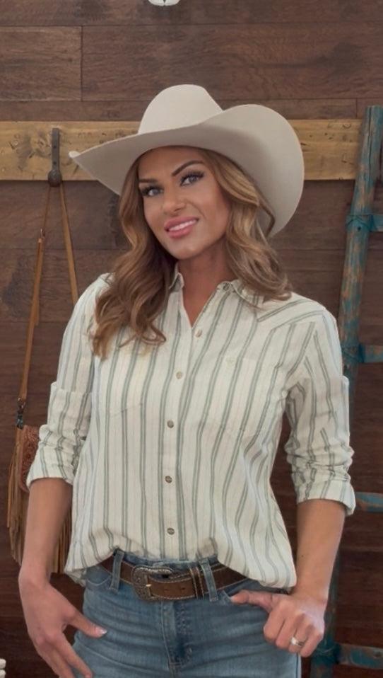 Woman wearing a cowboy hat, striped shirt, and jeans in a rustic setting.