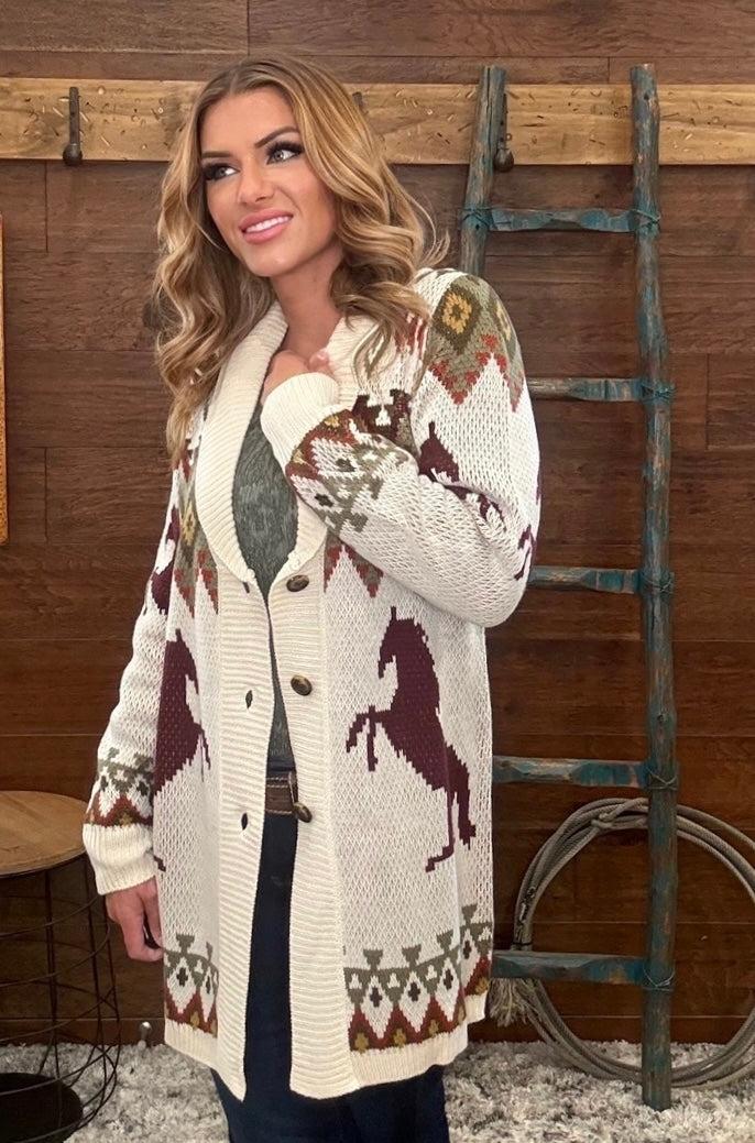 A woman wearing a cream knit cardigan with brown horse motifs and geometric patterns, standing in front of a rustic wooden wall with a ladder.