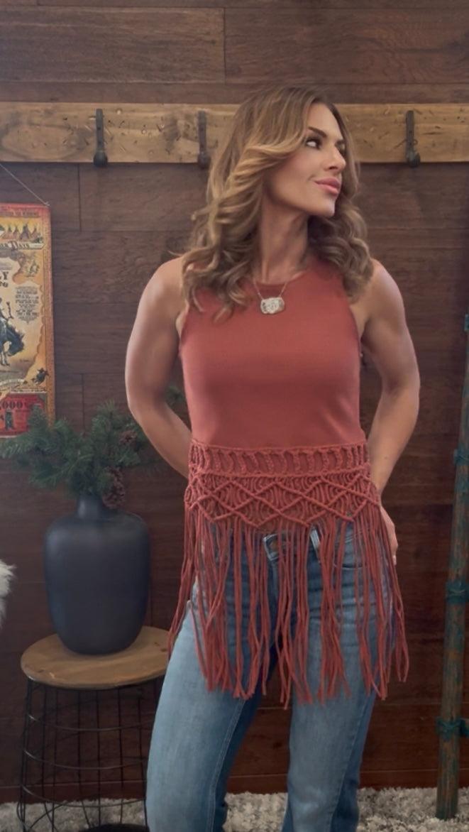 Woman wearing a rust-colored fringe top and jeans against a wooden wall.