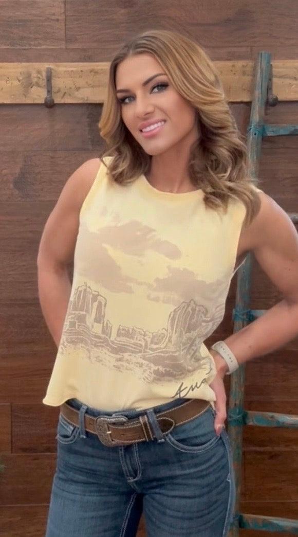 Woman wearing a yellow sleeveless top with a graphic design and blue jeans against a wooden background