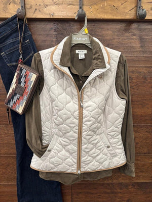A quilted sleeveless vest in off-white with tan trim, layered over an olive green button-up shirt, hanging on a gray ARIAT branded hanger against a dark wood background.
