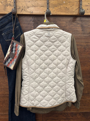 A quilted sleeveless vest in off-white with tan trim, layered over an olive green button-up shirt, hanging on a gray ARIAT branded hanger against a dark wood background.