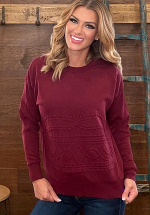 A smiling blonde woman wearing a long-sleeve crewneck sweater in burgundy with a textured knit pattern on the front, paired with blue jeans, standing against a rustic wooden wall.