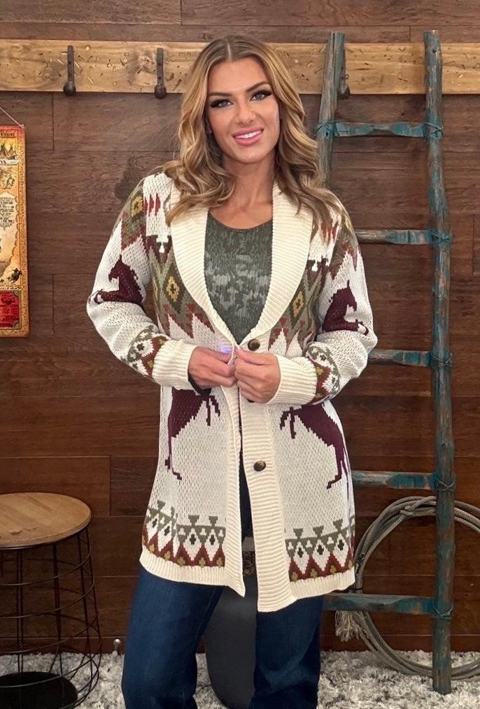 A woman modeling a cream-colored knit cardigan with brown and burgundy geometric patterns, styled over a dark gray top and blue jeans, standing in front of a rustic wooden wall with a ladder.