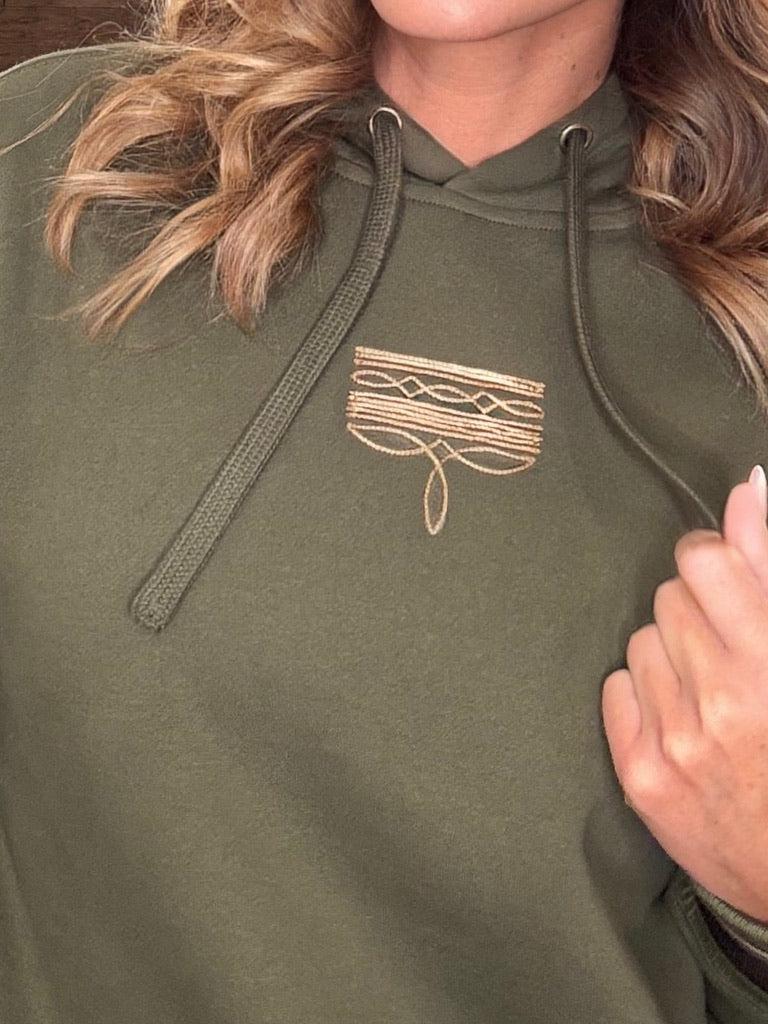 Close-up of a person wearing an olive green hoodie with a small gold embroidered chest graphic and matching drawstrings.