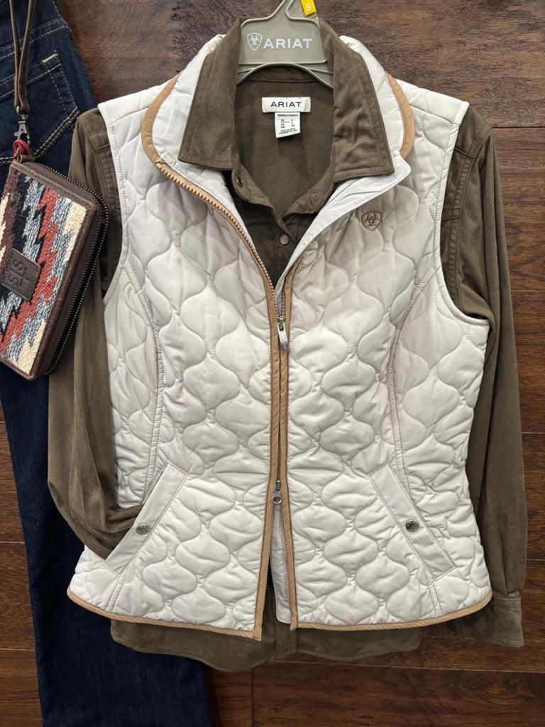 A quilted sleeveless vest in off-white with tan trim, layered over an olive green button-up shirt, hanging on a gray ARIAT branded hanger against a dark wood background.