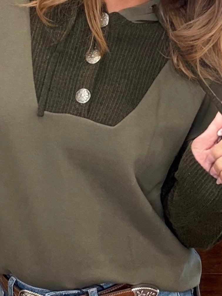 Close-up of a person wearing an olive green long-sleeve top with a dark olive ribbed yoke and silver buttons, paired with blue jeans and a brown belt.