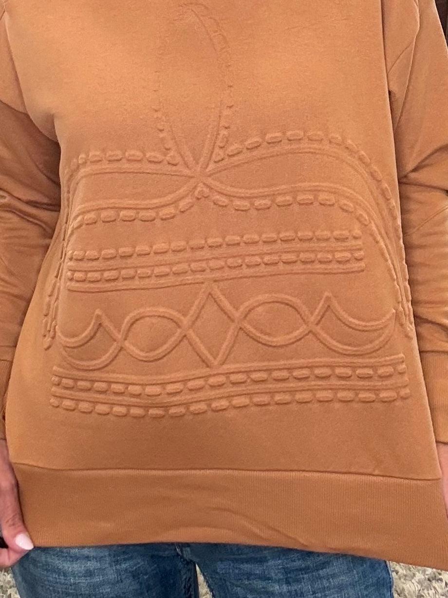 A close-up of a person wearing a camel brown long-sleeve knit sweater with a raised geometric pattern on the front, paired with blue jeans.