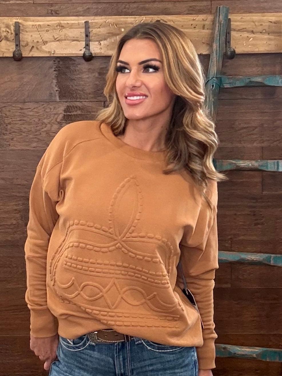 A woman wearing a camel-colored crewneck sweatshirt with a raised western-style pattern on the front, paired with blue jeans, standing against a rustic wooden wall.