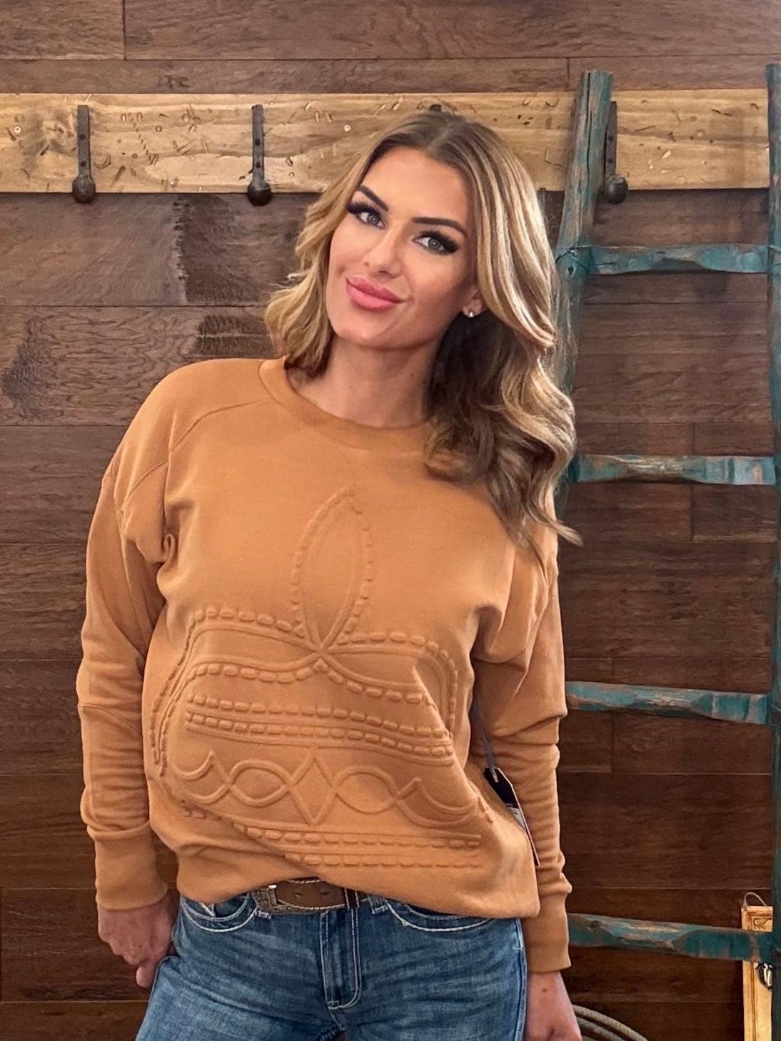 A woman wearing a camel-colored crewneck sweatshirt with a textured cable-knit bow design on the front, paired with blue jeans, standing against a rustic wooden wall with a teal ladder.