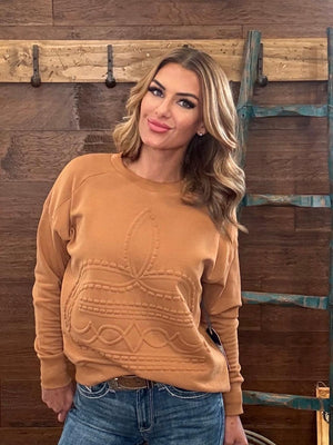 A woman wearing a camel-colored crewneck sweatshirt with a textured cable-knit bow design on the front, paired with blue jeans, standing against a rustic wooden wall with a teal ladder.
