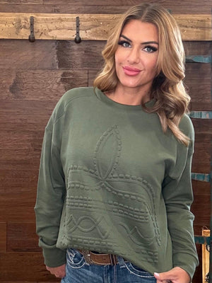 A woman wearing an olive green crewneck sweatshirt with a raised boot stitch pattern standing in front of a rustic wooden wall