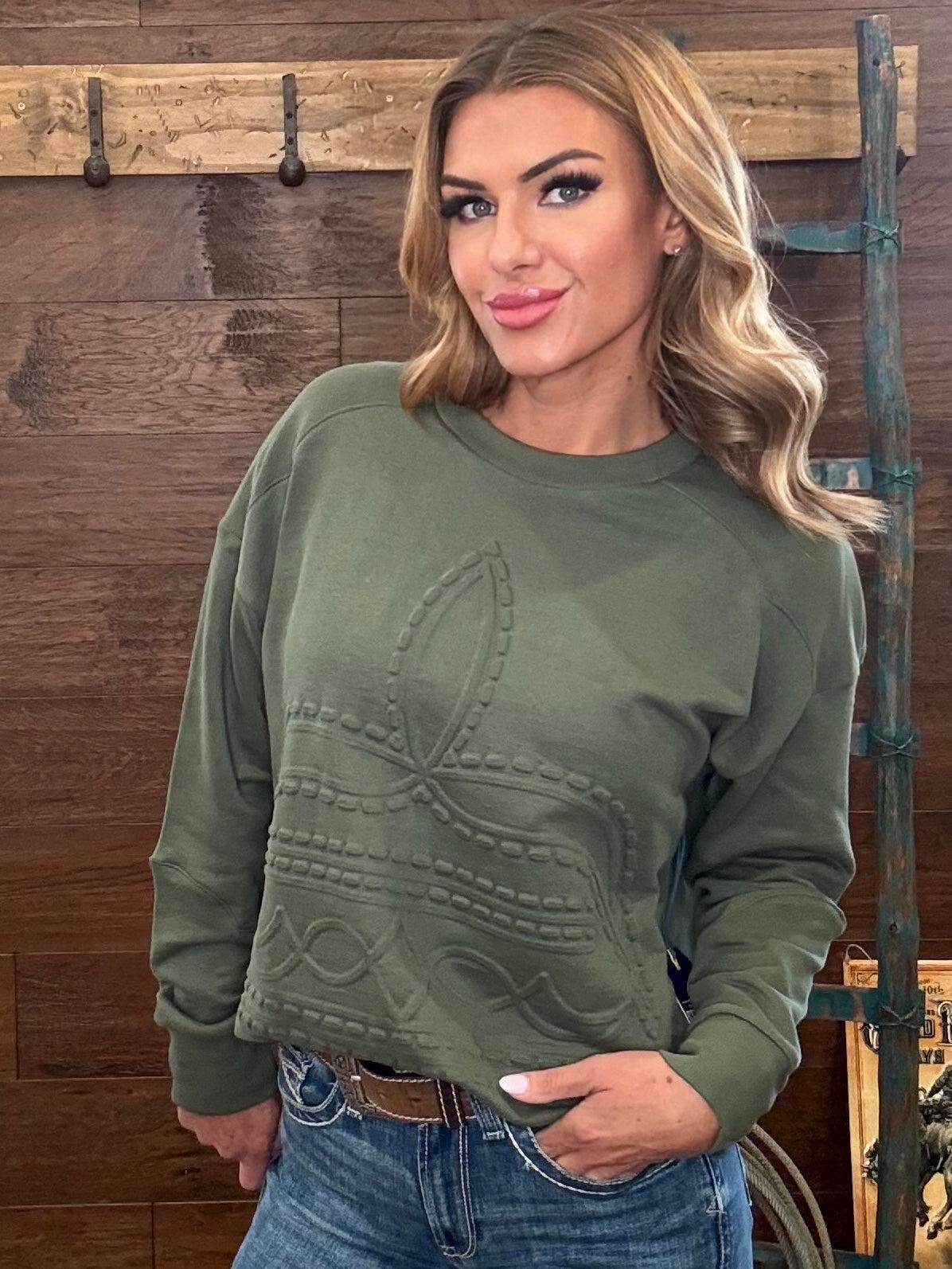 A woman wearing an olive green long-sleeve crewneck sweatshirt with a raised boot stitch pattern on the front, paired with blue jeans, standing against a rustic wooden wall.