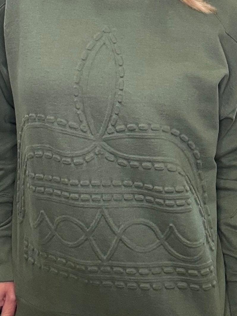Close-up of a person wearing an olive green long-sleeve top or sweatshirt with a large tonal embroidered or raised boot stitch pattern. 