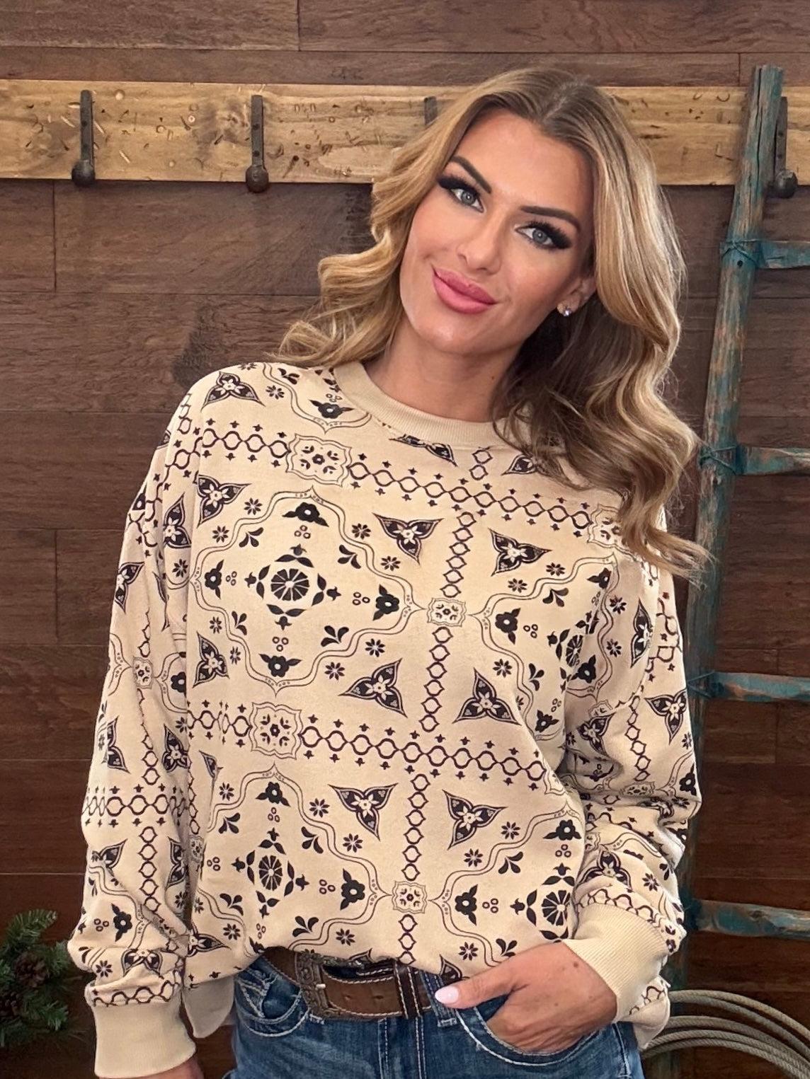 A woman wearing a beige long-sleeve crewneck sweatshirt with a black geometric tribal pattern, paired with blue jeans, standing in front of a rustic wooden wall.