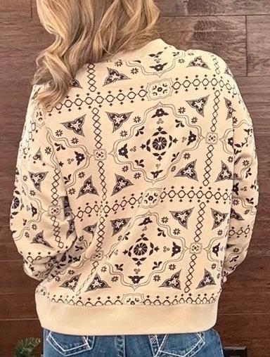 A person viewed from behind wearing a cream-colored bomber jacket with a black geometric and floral pattern, paired with blue jeans against a dark wood plank background.