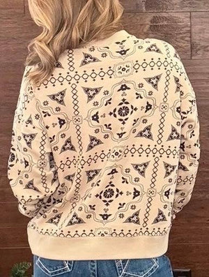 A person viewed from behind wearing a cream-colored bomber jacket with a black geometric and floral pattern, paired with blue jeans against a dark wood plank background.