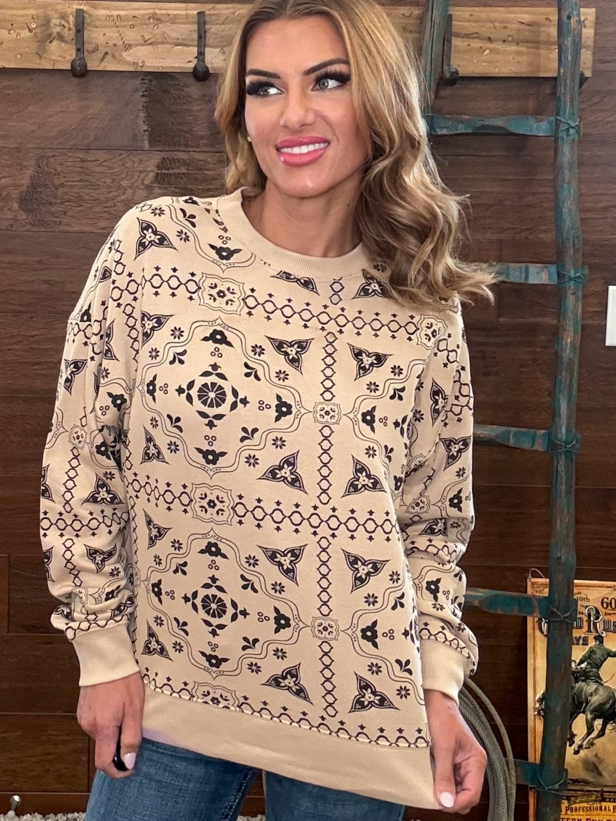 A woman wearing a beige long-sleeve crewneck sweatshirt with a black geometric tribal pattern, standing in front of a rustic wooden wall.