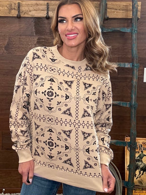 A woman wearing a beige long-sleeve crewneck sweatshirt with a black geometric tribal pattern, standing in front of a rustic wooden wall.