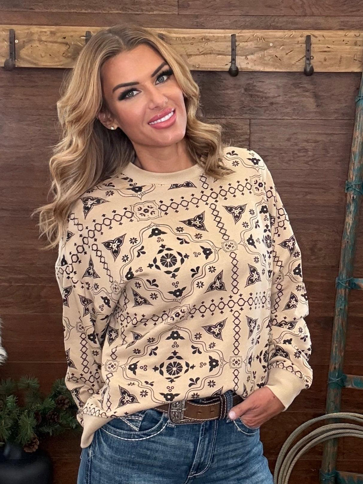 A woman wearing a beige long-sleeve crewneck sweatshirt with a black geometric southwestern-style pattern, paired with blue jeans and a brown belt, standing in front of a rustic wooden wall.