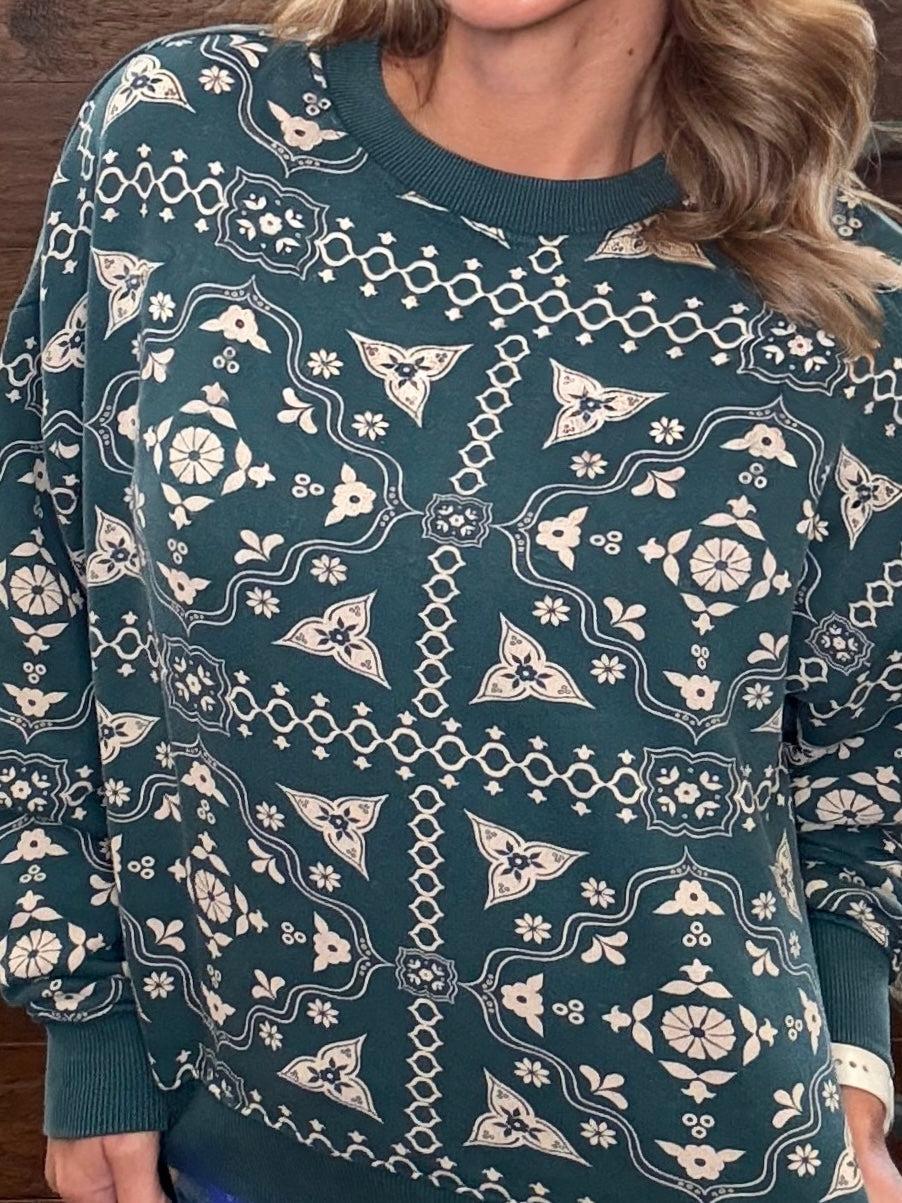 A close-up of a woman wearing a dark teal crewneck sweater with a cream-colored geometric and floral pattern.