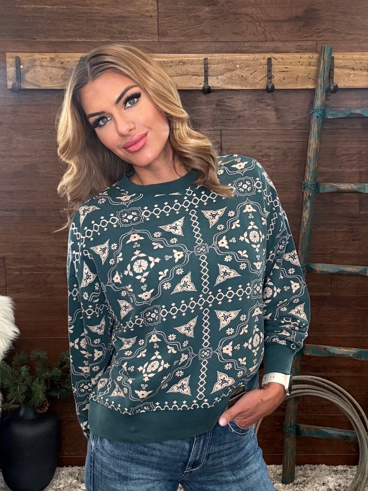 A woman wearing a dark teal green long-sleeve crewneck sweatshirt with a cream-colored geometric and floral pattern, paired with blue jeans, standing in front of a rustic wooden wall.