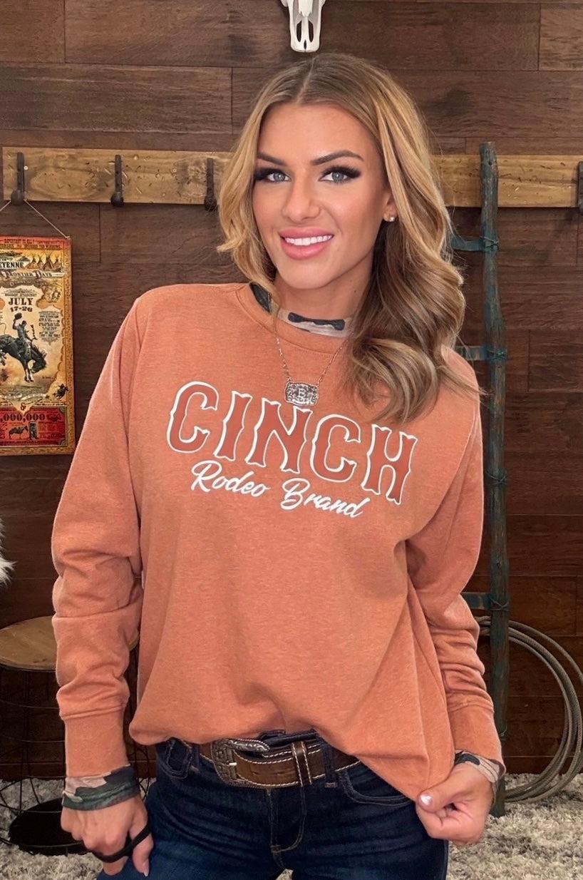 A woman wearing a burnt orange crewneck sweatshirt with 'CINCH Rodeo Brand' printed in white on the front, paired with dark blue jeans, standing in front of a rustic wooden wall.