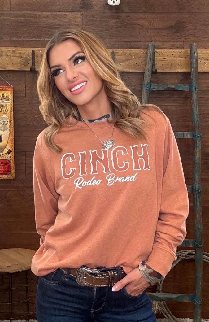 A woman wearing a terracotta-colored long-sleeve sweatshirt with 'CINCH Rodeo Brand' printed on the front, paired with dark blue jeans, standing in front of a rustic wooden wall.