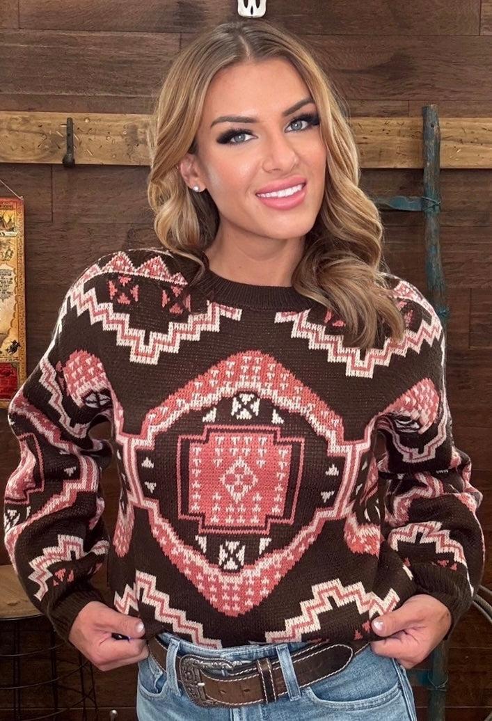 A woman wearing a brown knit sweater with a pink and white geometric southwestern pattern, paired with blue jeans, standing in front of a rustic wooden wall.