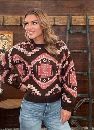 A woman wearing a brown knit sweater with pink geometric southwestern pattern, paired with light blue jeans, standing in front of a rustic wooden wall.