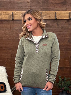 A woman wearing an olive green quarter-zip fleece pullover with a patterned collar and zipper trim, standing in front of a rustic wooden wall.