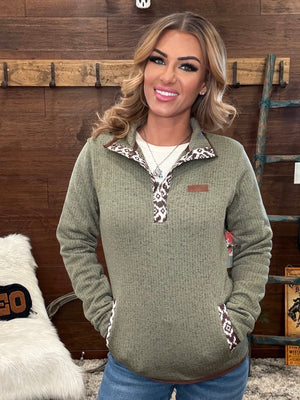 A woman wearing an olive green waffle-knit quarter-pullover with patterned trim on the collar and pockets, paired with blue jeans, standing in front of a rustic wood-paneled wall.