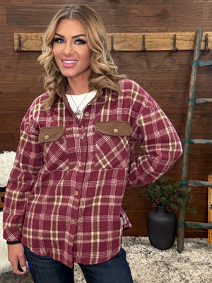 A woman wearing a long-sleeve button-up plaid shirt in burgundy and beige, with brown chest flap pockets, posed in front of a rustic wood wall with a ladder and potted plant.