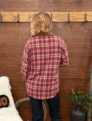 A person viewed from behind wearing a red and beige plaid flannel shirt and dark jeans, standing in front of a wooden wall with a coat rack.