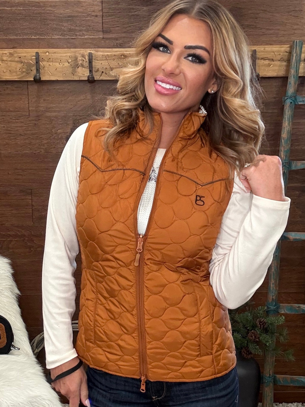 A woman wearing a quilted rust-orange sleeveless puffer vest over a white long-sleeve shirt and dark jeans, standing in front of a rustic wooden wall.