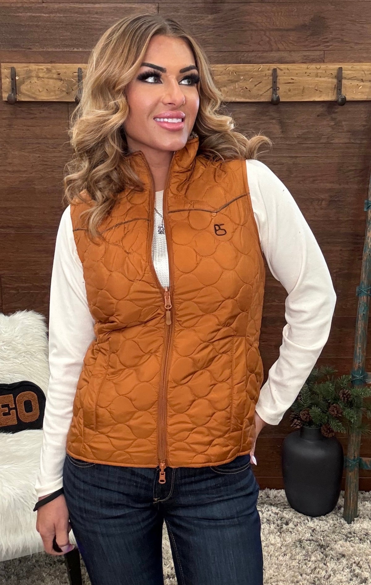 A woman wearing a quilted sleeveless zip-front vest in a warm camel brown color over a white long-sleeve shirt and dark blue jeans, standing in front of a rustic wooden wall.