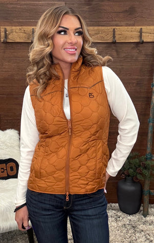 A woman wearing a quilted sleeveless zip-front vest in a warm camel brown color over a white long-sleeve shirt and dark blue jeans, standing in front of a rustic wooden wall.