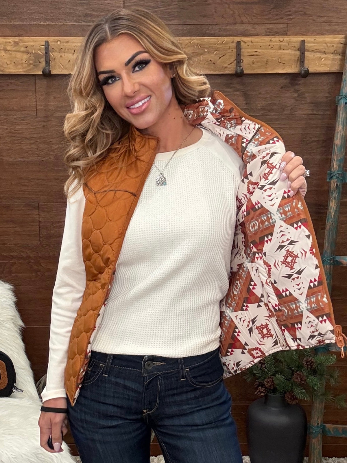 A woman models a split-design vest with a quilted rust-orange front panel and a patterned winter-themed lining featuring snowflakes and geometric motifs in red, white, and brown, worn over a cream ribbed knit sweater and dark blue jeans.