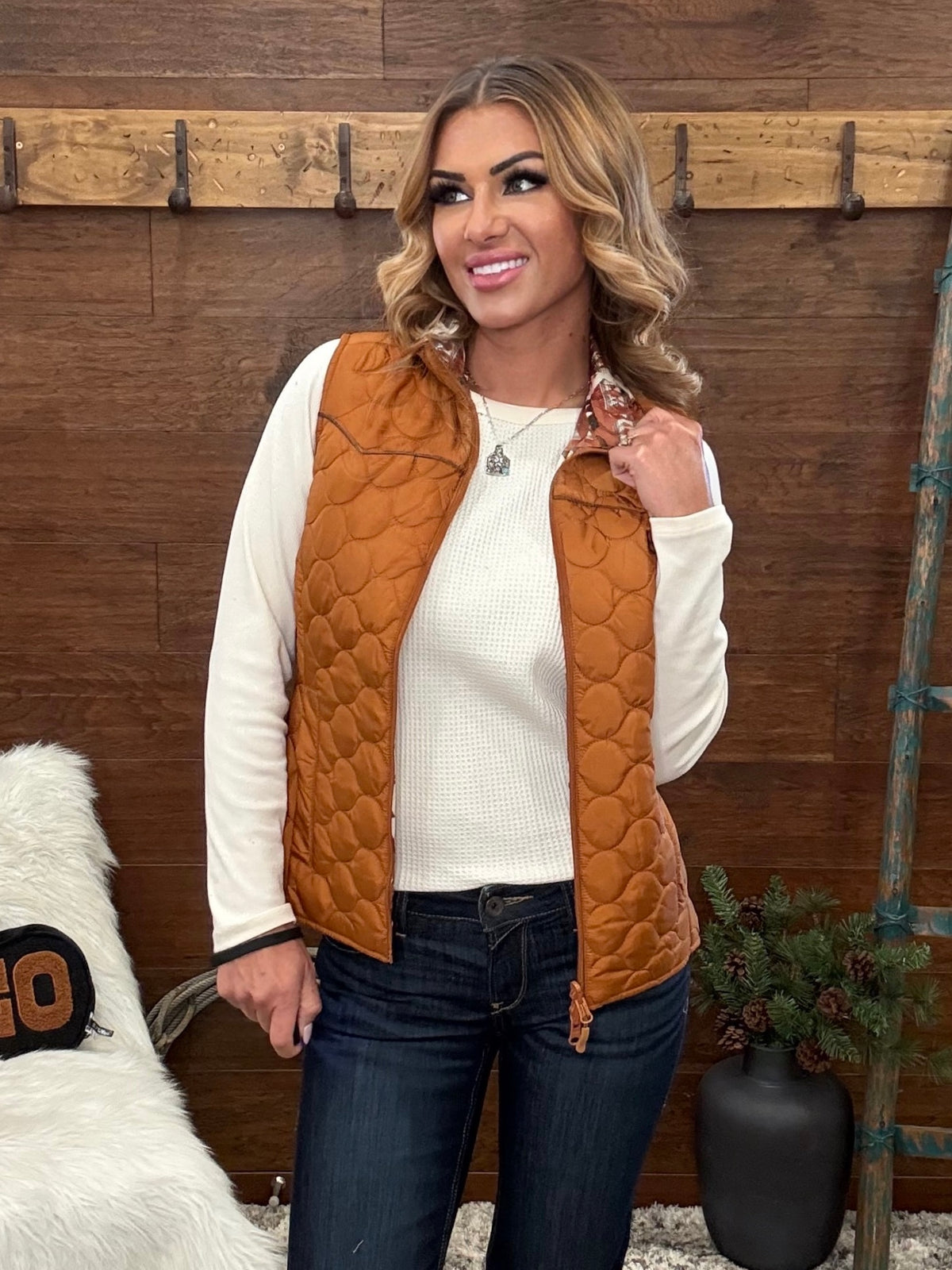 A woman wearing a quilted rust-orange zip-front vest over a cream ribbed long-sleeve top and dark blue jeans, standing in front of a rustic wooden wall.
