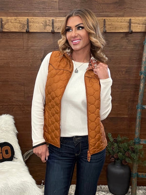 A woman wearing a quilted rust-orange zip-front vest over a cream ribbed long-sleeve top and dark blue jeans, standing in front of a rustic wooden wall.