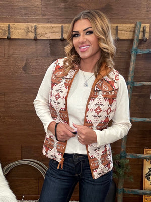 A woman wearing a sleeveless western-style vest with a brown and rust geometric pattern over a cream knit top and dark blue jeans, standing in front of a rustic wood-paneled wall.