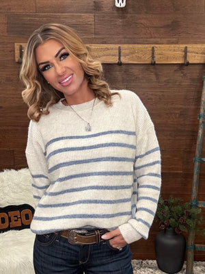 A woman wearing a cream knit sweater with horizontal blue stripes, paired with dark blue jeans and a brown belt, standing in front of a rustic wooden wall.