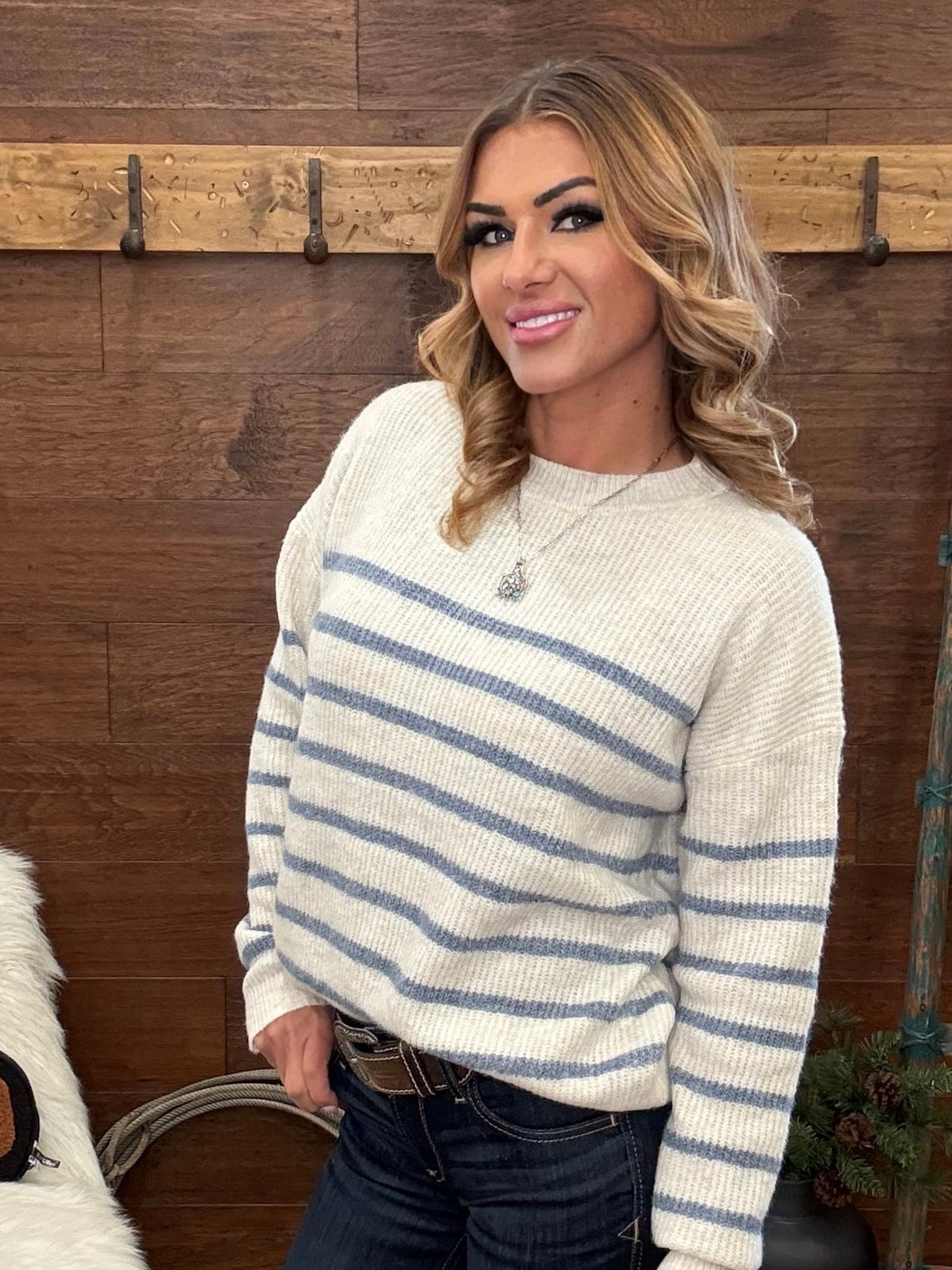 A woman wearing a cream-colored ribbed knit sweater with horizontal blue-gray stripes, paired with dark blue jeans, standing in front of a rustic wooden wall.